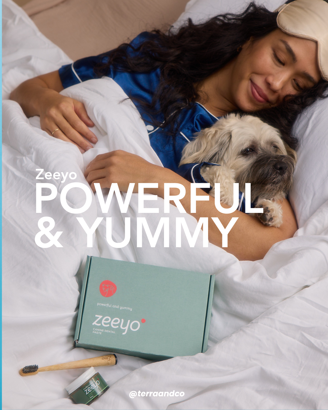 A cozy lifestyle photo of a woman in blue silk pajamas cuddling her small scruffy dog in white bedding. In the foreground, a Zeeyo 'powerful and yummy' canine dental kit, bamboo toothbrush, and glass jar of toothpaste are nestled in the blankets, symbolizing a gentle and healthy bedtime routine.