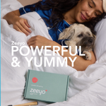 A cozy lifestyle photo of a woman in blue silk pajamas cuddling her small scruffy dog in white bedding. In the foreground, a Zeeyo 'powerful and yummy' canine dental kit, bamboo toothbrush, and glass jar of toothpaste are nestled in the blankets, symbolizing a gentle and healthy bedtime routine.