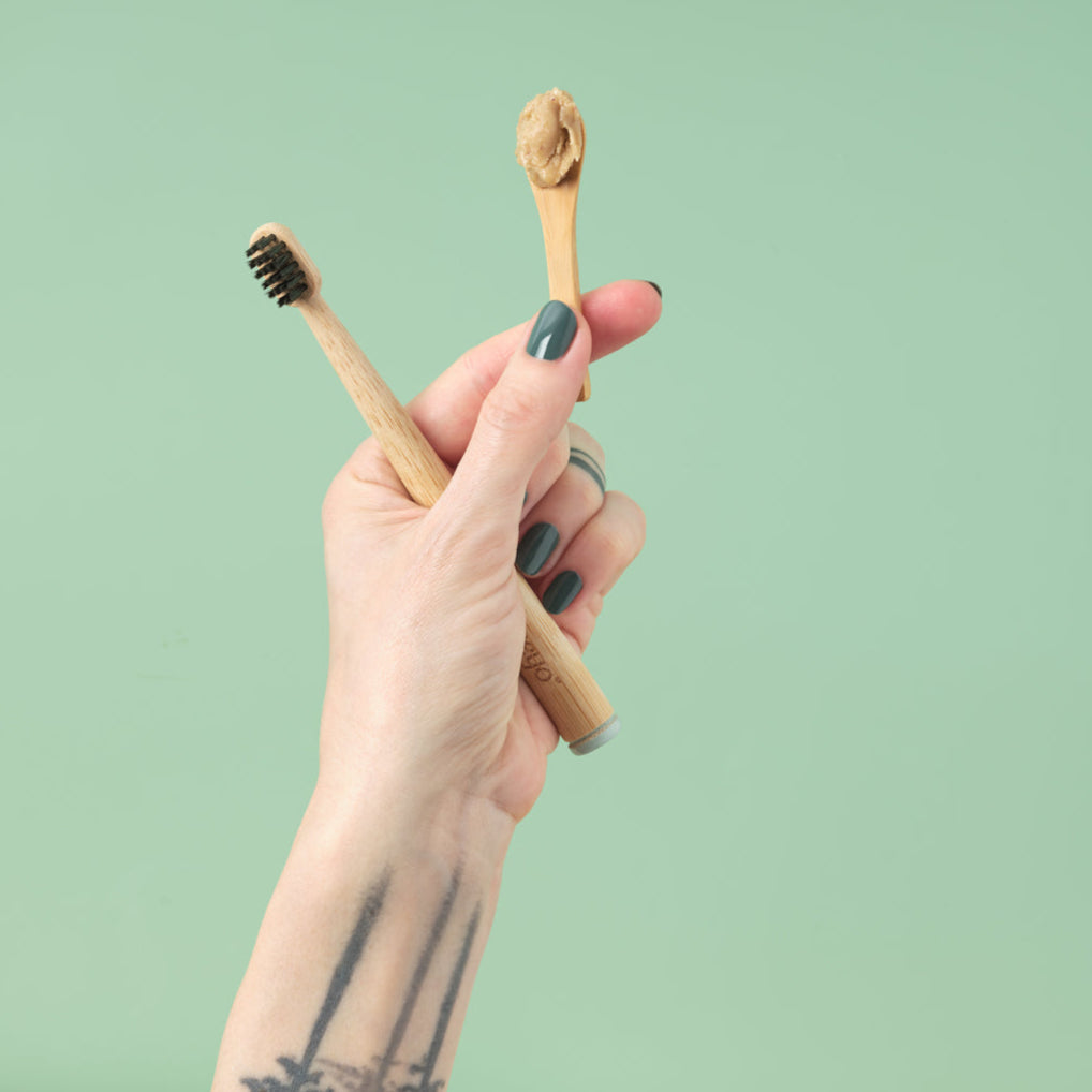 A close-up view of a hand holding the Zeeyo sustainable bamboo-handled dog toothbrush and a separate bamboo scoop displaying a single dollop of natural, hydroxyapatite-rich doggie toothpaste against a soft sage-green background.