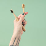 A close-up view of a hand holding the Zeeyo sustainable bamboo-handled dog toothbrush and a separate bamboo scoop displaying a single dollop of natural, hydroxyapatite-rich doggie toothpaste against a soft sage-green background.