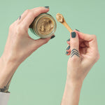 A top-down view of a person’s hands holding an open glass jar of Zeeyo dog toothpaste and a small bamboo scoop with a sample of the creamy, thick, and natural-colored dental paste against a sage green background.